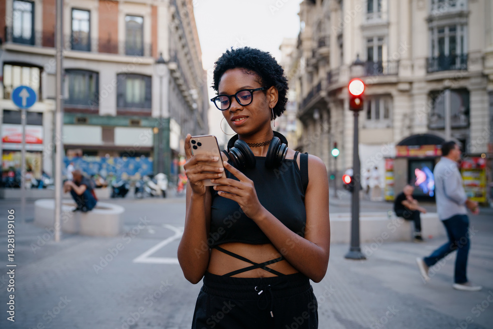 Young Black woman using smartphone outdoors in city during daytime, headphones resting on neck, checking playlist or messages with a calm and focused expression