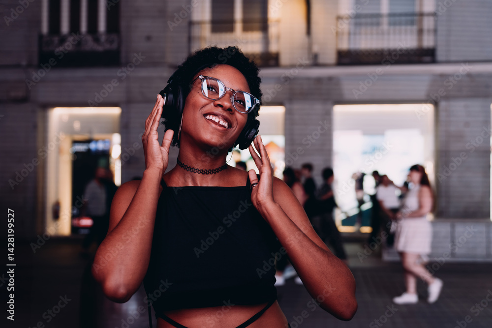 Happy young Black woman enjoying music in headphones while standing on lively night street, expressing joy through sound, movement, and personal style