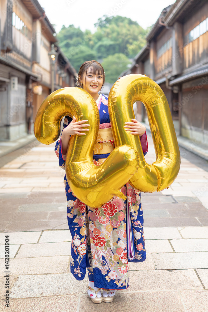 On a summer day, a 20-year-old Japanese girl wearing a blue kimono is in the traditional townscape of a Japanese city. She is holding large golden balloons shaped like the numbers 2 and 0.