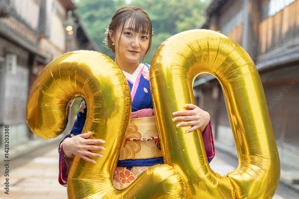 On a summer day, a 20-year-old Japanese girl wearing a blue kimono is in the traditional townscape of a Japanese city. She is holding large golden balloons shaped like the numbers 2 and 0.