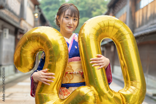 Obraz na płótnie On a summer day, a 20-year-old Japanese girl wearing a blue kimono is in the traditional townscape of a Japanese city. She is holding large golden balloons shaped like the numbers 2 and 0. w salonie