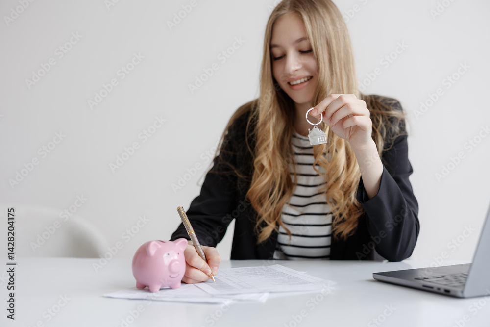 Young woman completing paperwork for new home purchase while holding house key and sitting next to piggy bank