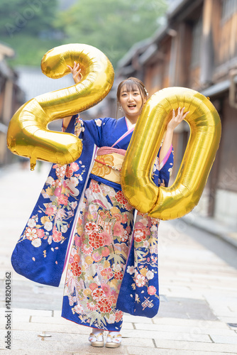 Obraz na płótnie On a summer day, a 20-year-old Japanese girl wearing a blue kimono is in the traditional townscape of a Japanese city. She is holding large golden balloons shaped like the numbers 2 and 0. w salonie