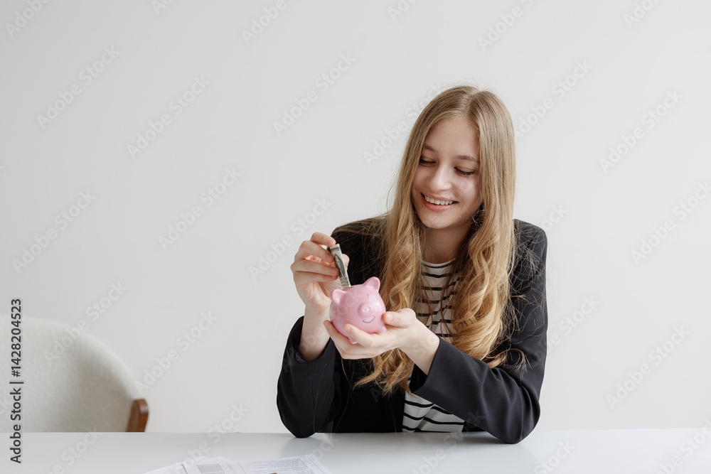 Young woman saving money in a pink piggy bank while sitting at a white desk