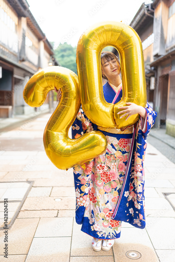 On a summer day, a 20-year-old Japanese girl wearing a blue kimono is in the traditional townscape of a Japanese city. She is holding large golden balloons shaped like the numbers 2 and 0.