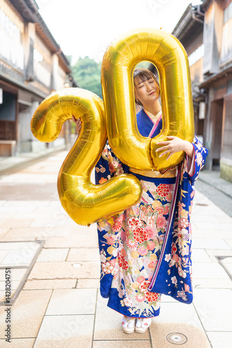 Obraz na płótnie On a summer day, a 20-year-old Japanese girl wearing a blue kimono is in the traditional townscape of a Japanese city. She is holding large golden balloons shaped like the numbers 2 and 0. w salonie