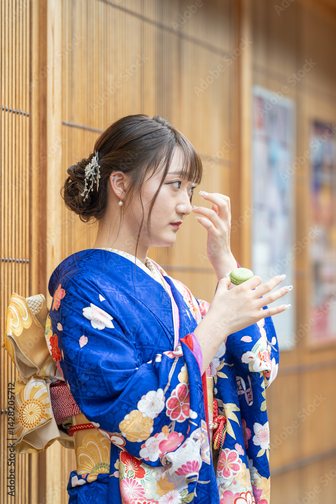 On a summer day, a 20-year-old Japanese girl wearing a blue kimono is standing in the traditional townscape of a Japanese city, holding a green macaron.