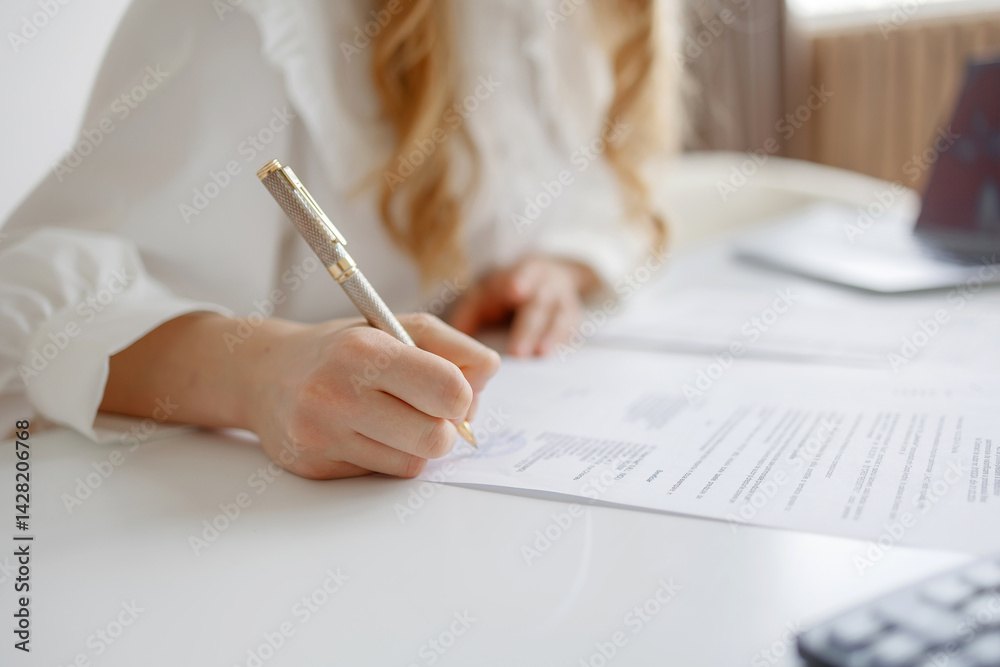 Woman writing on documents at a white desk with a calculator and office supplies in a well-lit workspace