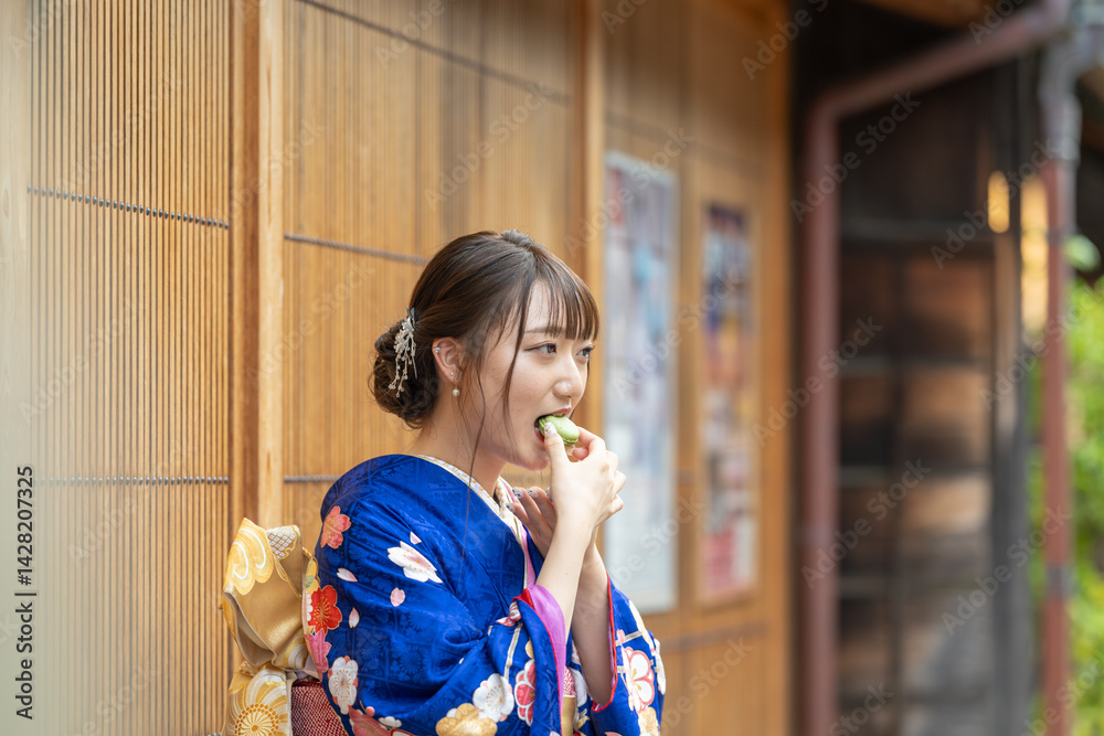 On a summer day, a 20-year-old Japanese girl wearing a blue kimono is standing in the traditional townscape of a Japanese city, eating a green macaron.