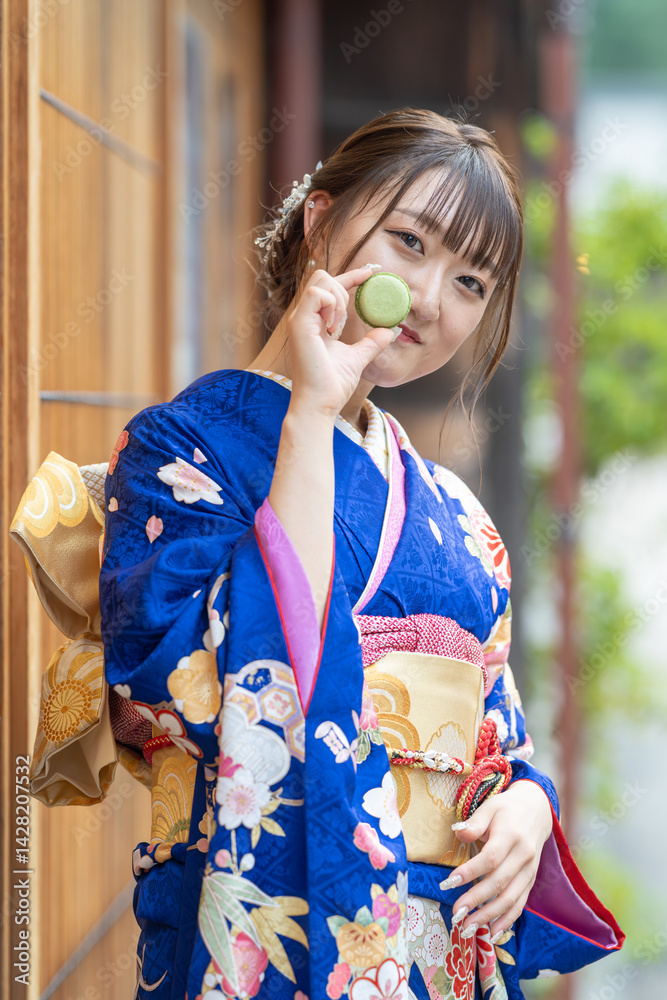 On a summer day, a 20-year-old Japanese girl wearing a blue kimono is standing in the traditional townscape of a Japanese city, holding a green macaron.