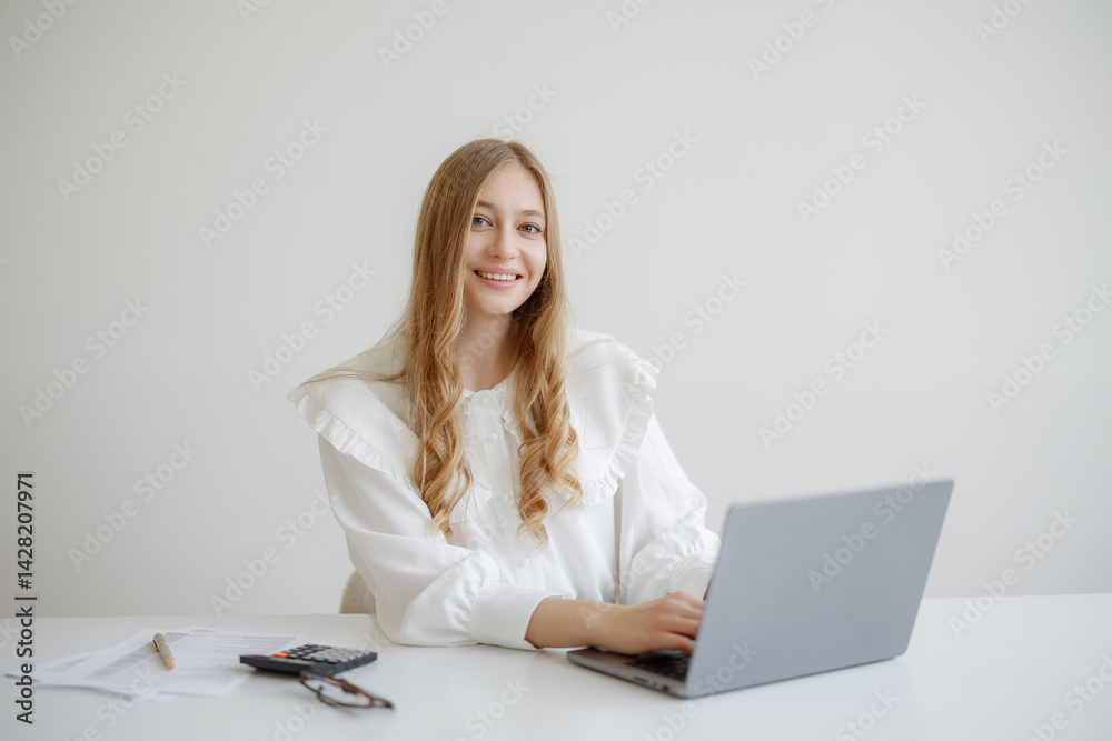 Young woman working on a laptop in a bright, minimal office while smiling towards the camera