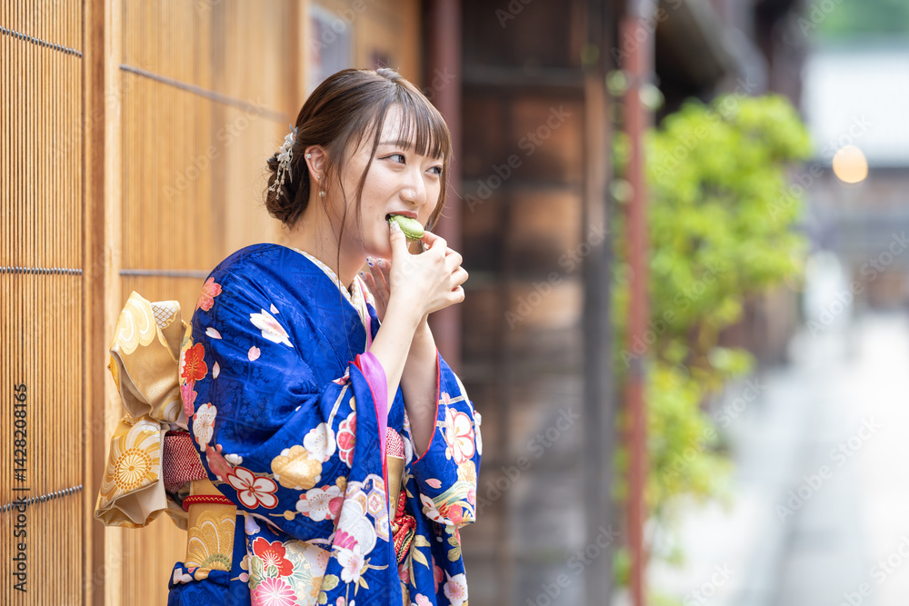 On a summer day, a 20-year-old Japanese girl wearing a blue kimono is standing in the traditional townscape of a Japanese city, eating a green macaron.