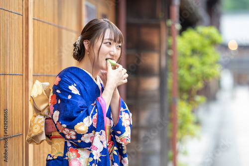 On a summer day, a 20-year-old Japanese girl wearing a blue kimono is standing in the traditional townscape of a Japanese city, eating a green macaron.