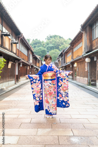 A teenage Japanese girl wearing a blue kimono stands in the view of a traditional town in a local Japanese city on a rainy summer day.