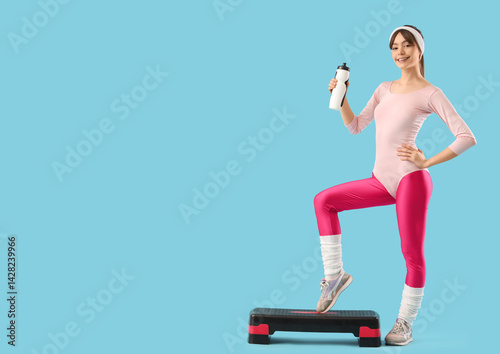 Sporty young woman with water bottle exercising on aerobic stepper against blue background