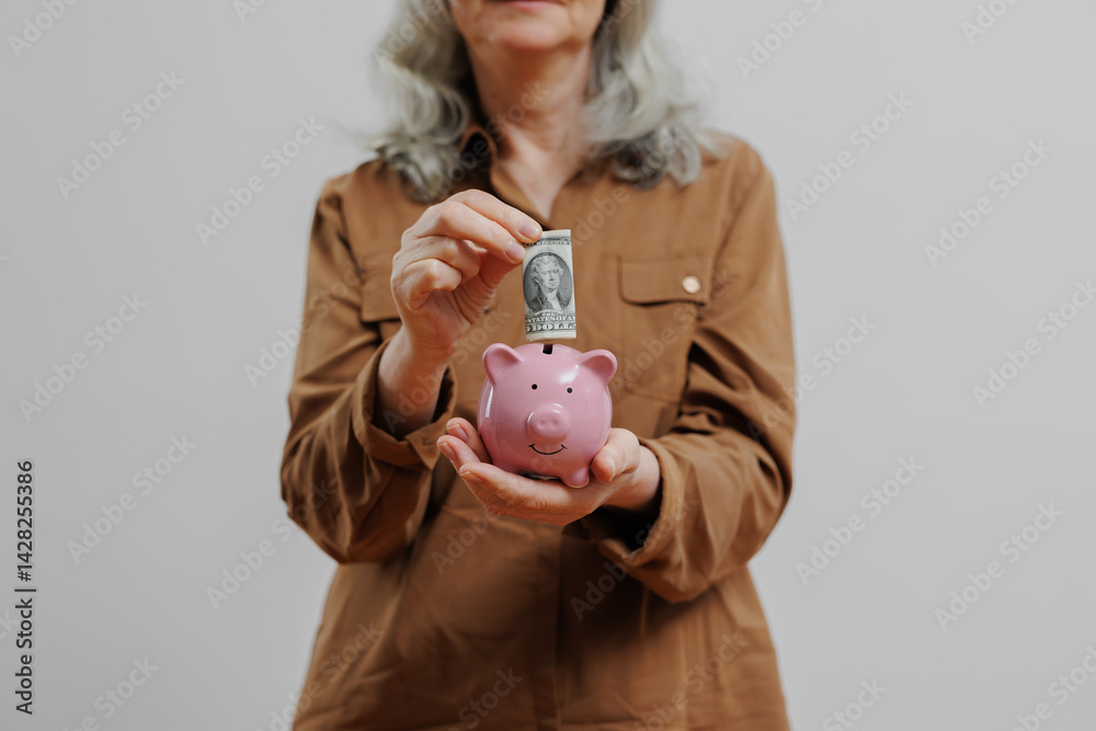 Senior woman depositing money into a pink piggy bank in a simple indoor setting while wearing a brown jacket