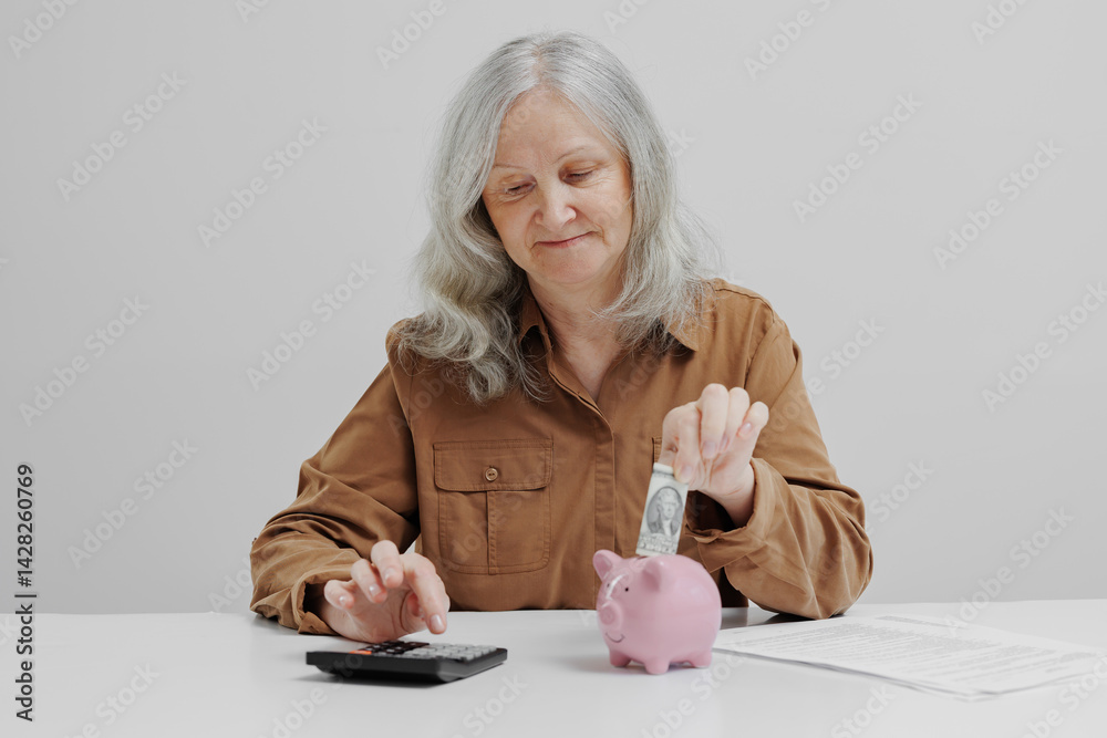 Elderly woman saving money in piggy bank while using calculator for budgeting in a minimalistic environment