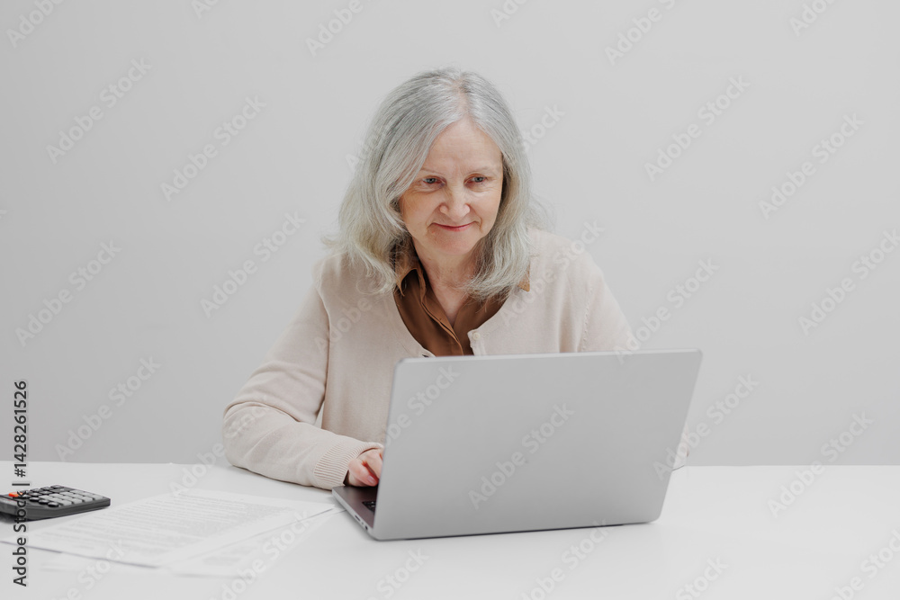 Senior woman using laptop while sitting at a desk with documents and a calculator in a modern office environment