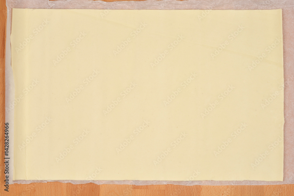A sheet of store-bought pale yellow puff pastry sits unfolded on parchment paper atop a wooden surface