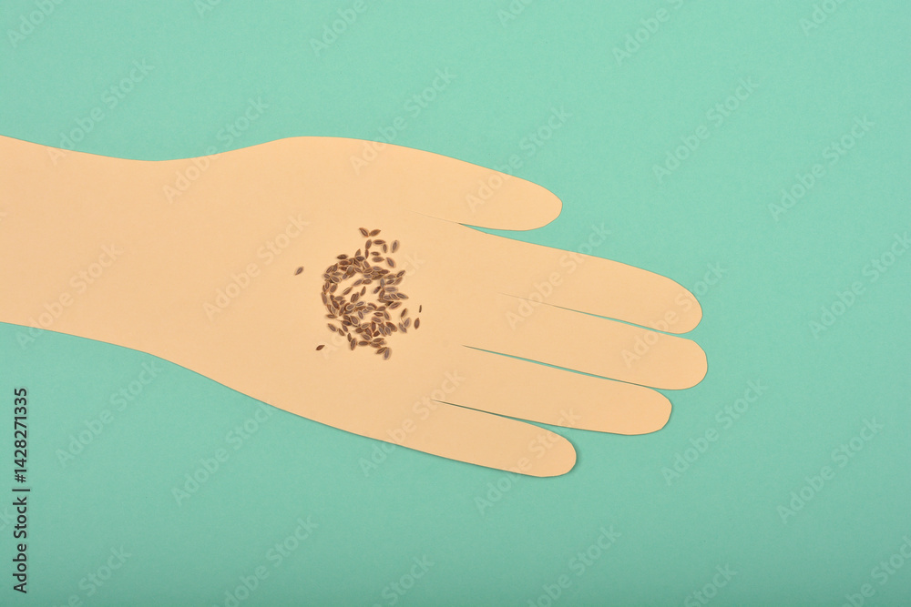 A top-down shot of dill (Anethum graveolens) seeds resting on a paper cutout hand to demonstrate size, against a teal background