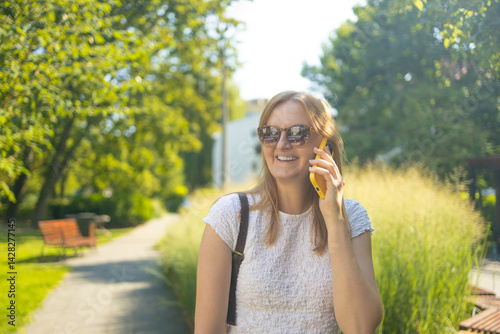 Obraz na płótnie Portrait of smiling 30s student freelancer woman looking at the camera talking on phone in summer park. People urban lifestyle concept. High quality photo w salonie
