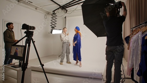 Young adult woman spending time at studio, woman model in fancy outfit taking part in a photoshoot in professional studio with photographer and talking with assistant.