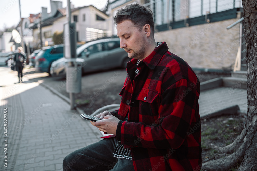 Male student with smartphone wearing plaid shirt sitting on bench while waiting for transport at tram or bus station. City public transport infrastructure, citizen and tourists transportation concept