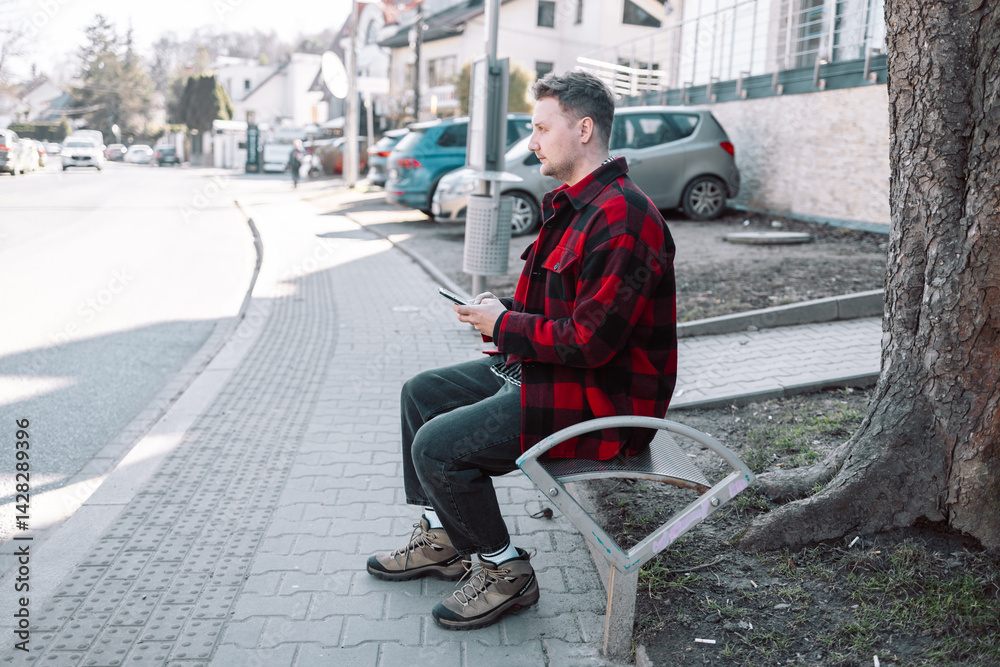 Male student with smartphone wearing plaid shirt sitting on bench while waiting for transport at tram or bus station. City public transport infrastructure, citizen and tourists transportation concept