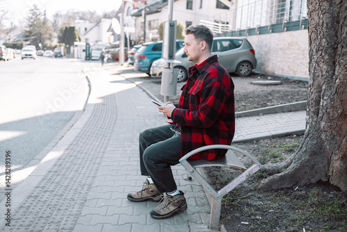 Obraz na płótnie Male student with smartphone wearing plaid shirt sitting on bench while waiting for transport at tram or bus station. City public transport infrastructure, citizen and tourists transportation concept w salonie