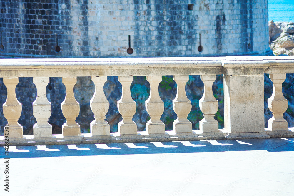 Stone balustrade with intricately carved balusters stands in foreground, casting shadows on tiled surface. Behind it weathered stone wall and a glimpse of the sea suggest a historic coastal location