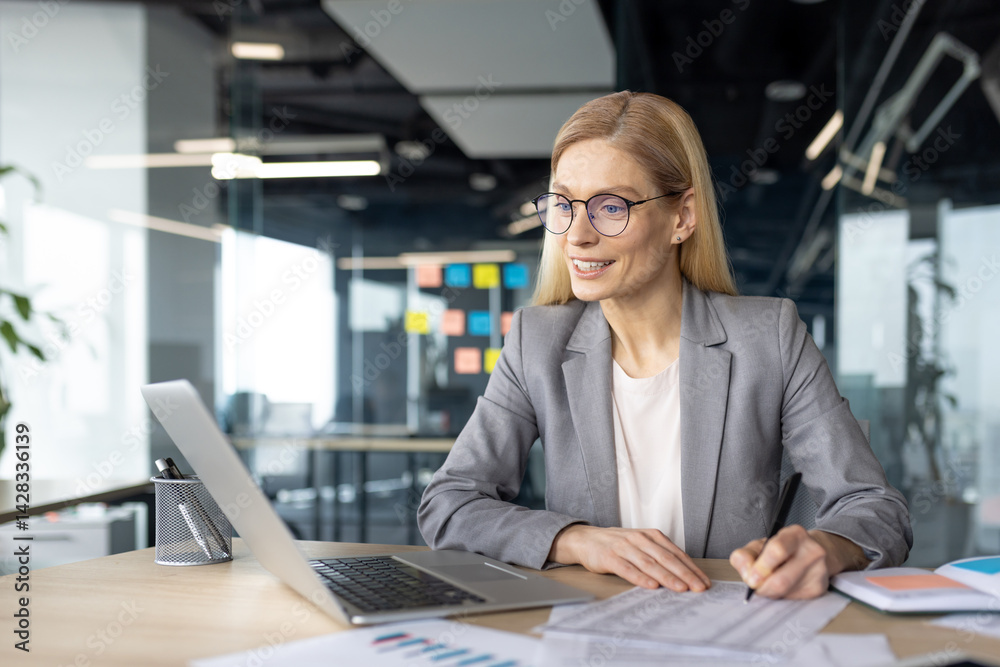 Obraz premium A professional woman in a gray suit works at her desk, focused on paperwork and her laptop in a modern office setting.