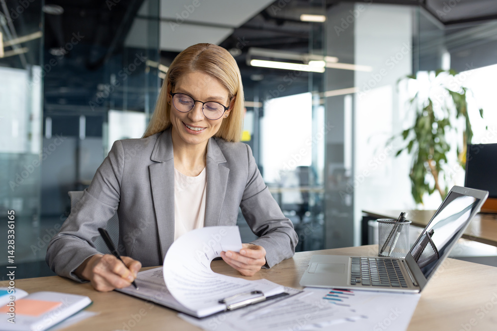 A smiling businesswoman in glasses meticulously reviewing documents while working in a modern office environment. She is wearing a stylish gray suit.