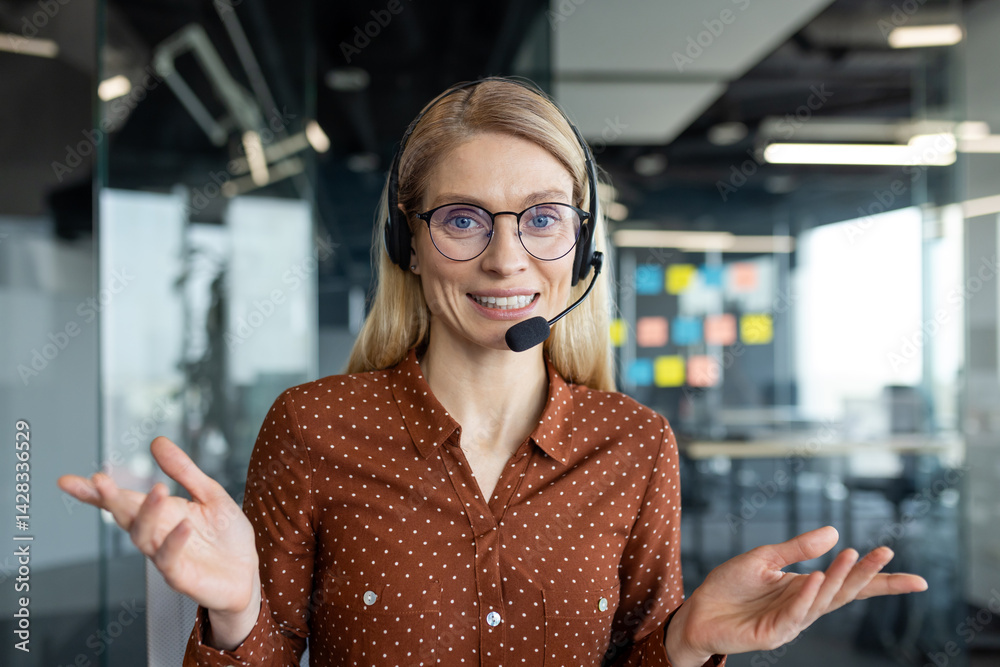 A friendly woman with glasses and a headset smiles while in an office, ready to assist. She gestures with her hands, showing openness and approachability.