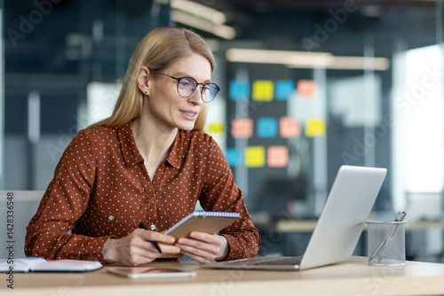 Obraz na płótnie A businesswoman is working on a laptop at her desk, holding a notebook, looking focused. She wears glasses in a modern office setting. w salonie