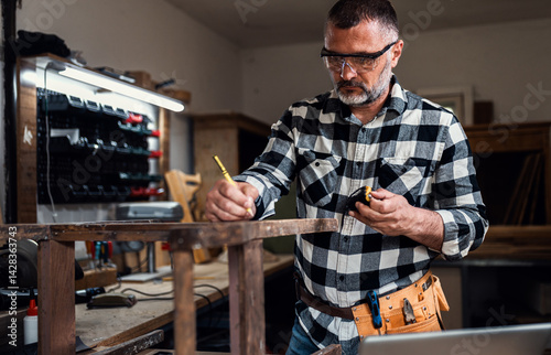 Carpenter standing in his workshop restoring old piece of furniture marking measurements on wood while using a laptop.
