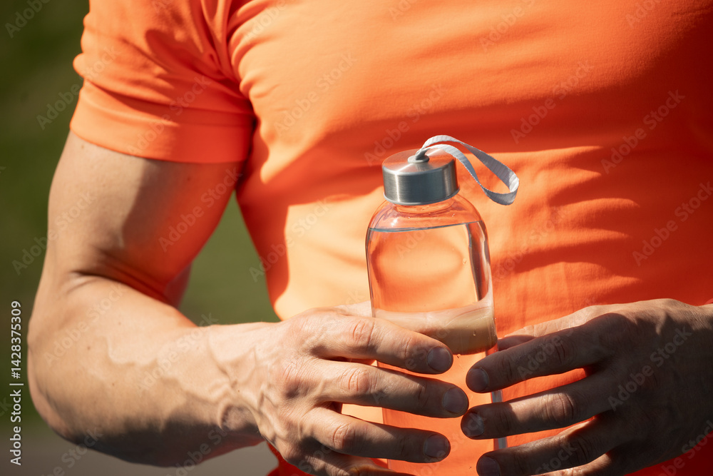 Muscular man holding water bottle in orange t-shirt on outdoor sports ground. Concept water balance for hot season for workout. Close up.