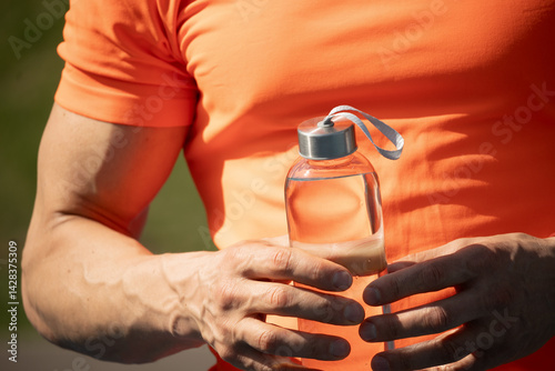 Obraz na płótnie Muscular man holding water bottle in orange t-shirt on outdoor sports ground. Concept water balance for hot season for workout. Close up. w salonie