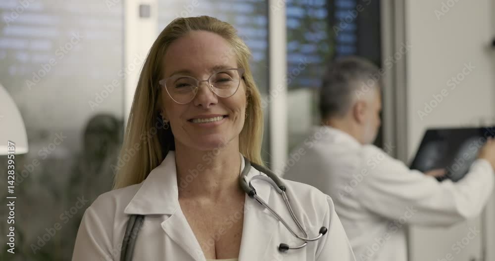 Smiling female doctor with stethoscope looks at camera in professional medical office as male colleague reviews brain scan in background Smiling female doctor with stethoscope looks at camera in professional medical office as male colleague reviews brain scan in background