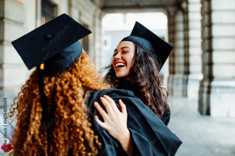 Two female students hugging each other, wearing graduation gown and cap, laughing and celebrating graduation day