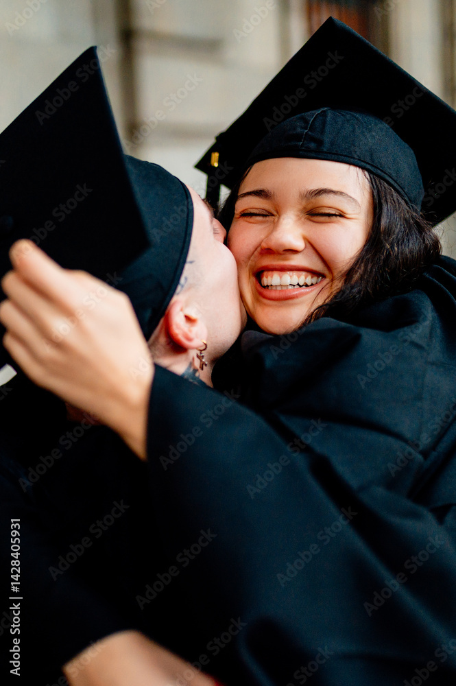 Two happy students wearing graduation gowns and caps are kissing and hugging each other on graduation day