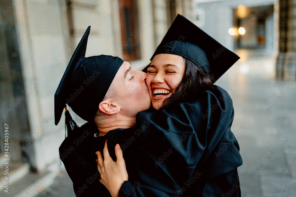 Two happy students wearing graduation gowns and caps are kissing and hugging on graduation day