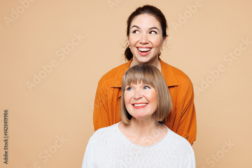 Elder parent mom 50s years old with young adult daughter two women together wearing casual clothes stand behind mother look aside on area isolated on plain pastel beige background. Family day concept. photo