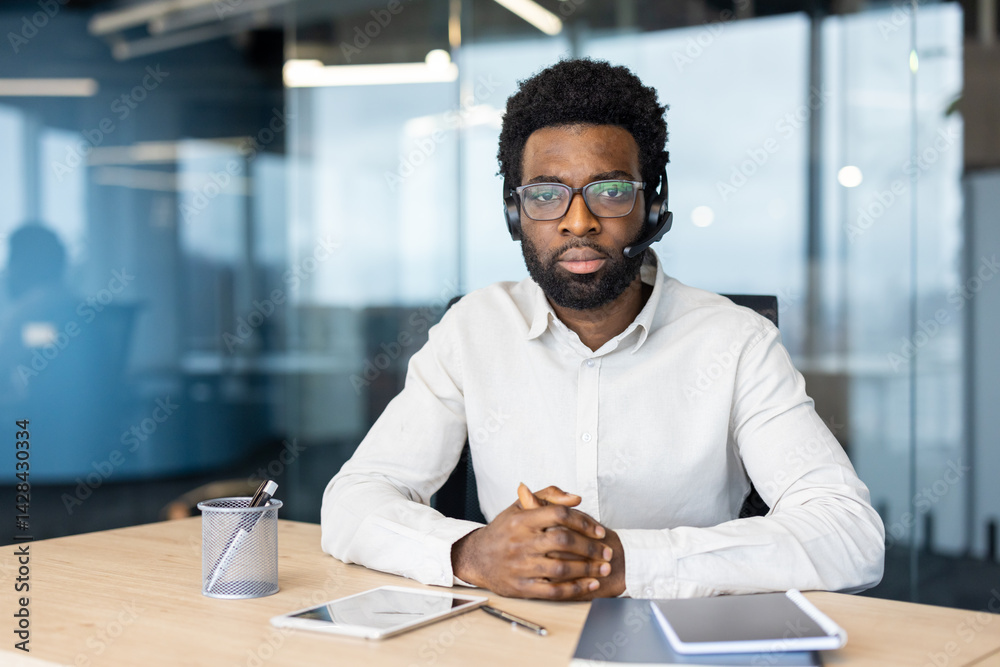 An african-american man wearing a headset sits in an office setting, looking at the camera with a serious expression, ready to assist.