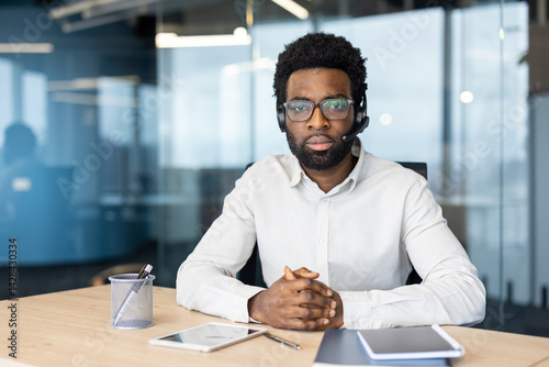 An african-american man wearing a headset sits in an office setting, looking at the camera with a serious expression, ready to assist.