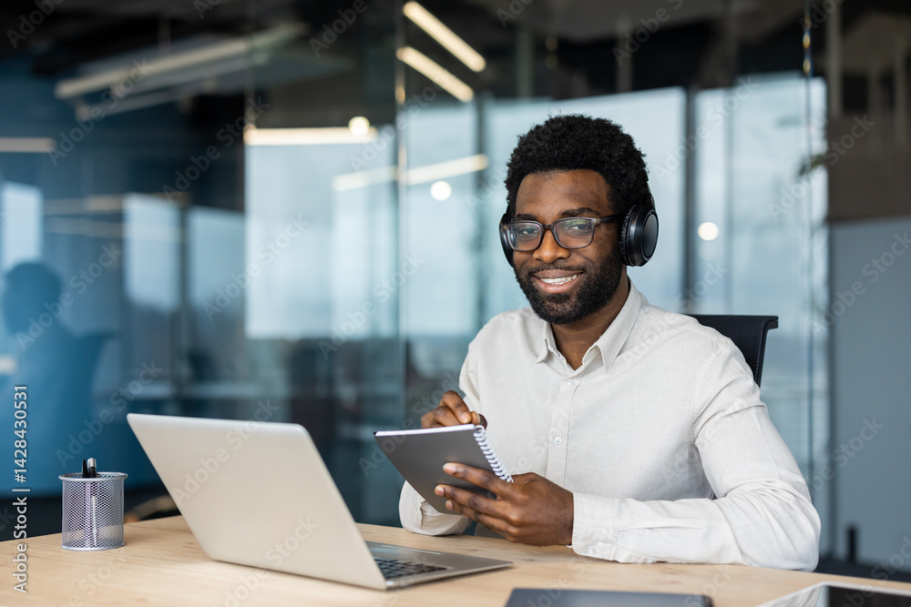 A smiling man is shown wearing headphones and glasses, working in an office setting at his desk, looking at the camera.