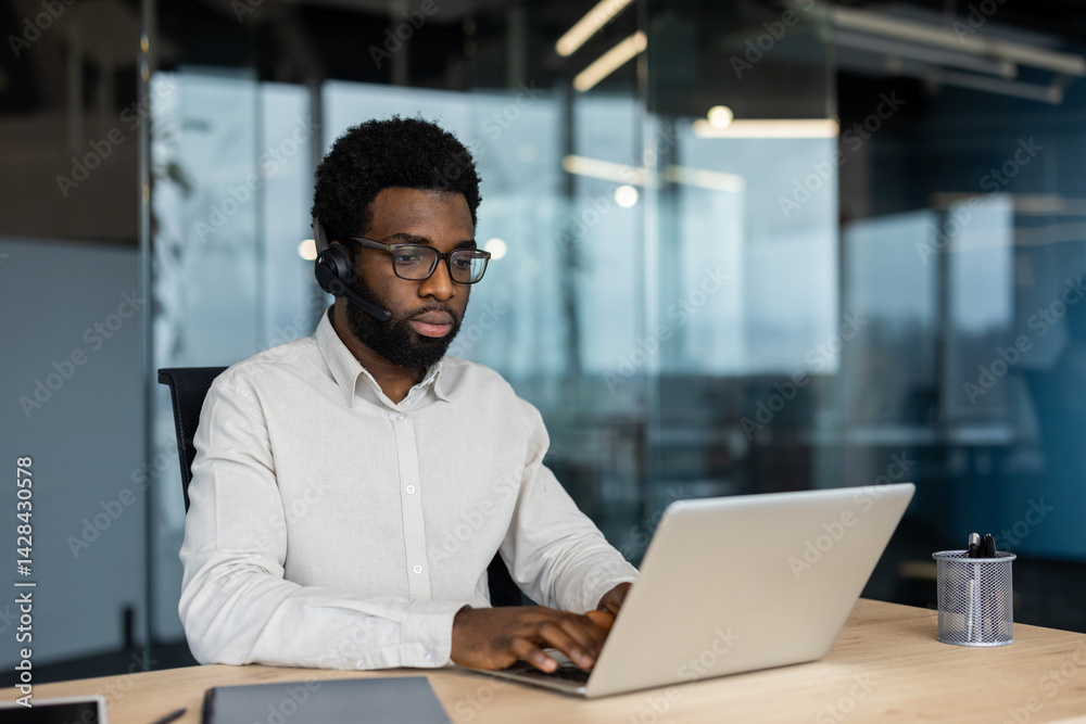 An african-american man wears a headset while working on his laptop in a modern office environment, focused on his tasks.