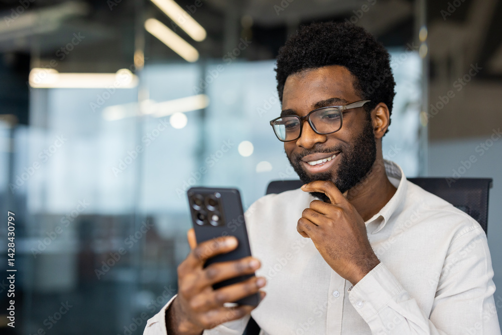 A handsome man in glasses smiles while looking at his phone in an office setting. He appears relaxed and is enjoying the moment.