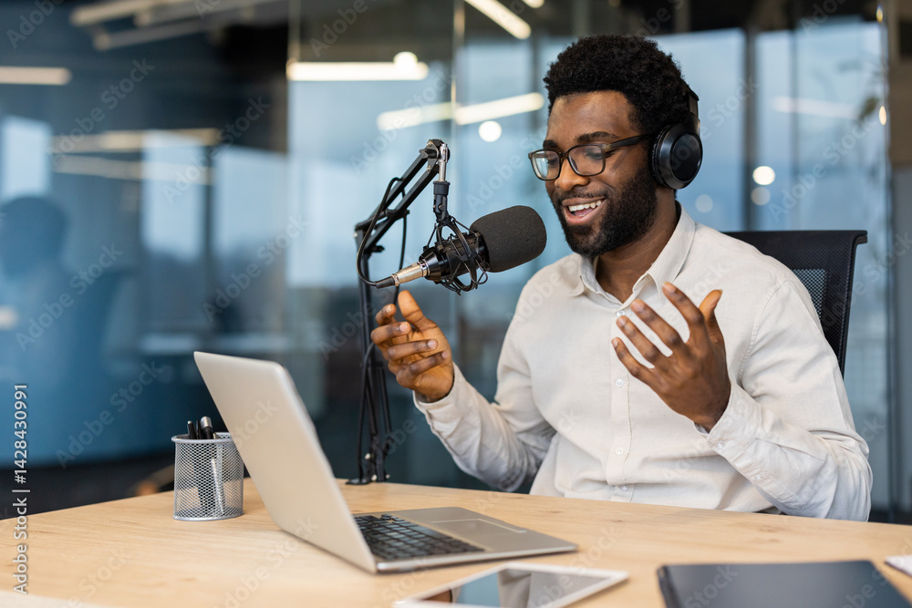 A smiling African-American man with glasses records a podcast in a modern office setting. He is using a microphone, headphones, and a laptop.