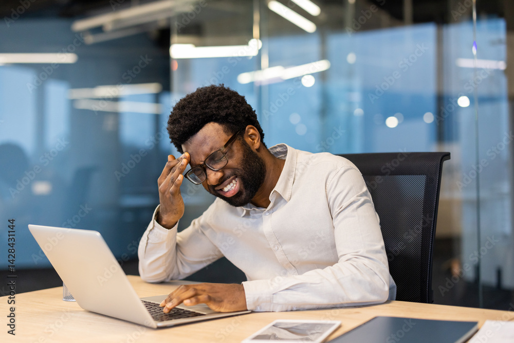 An African-American man working at his desk is shown with a stressed expression. He is experiencing a headache and appears overwhelmed while using his laptop.