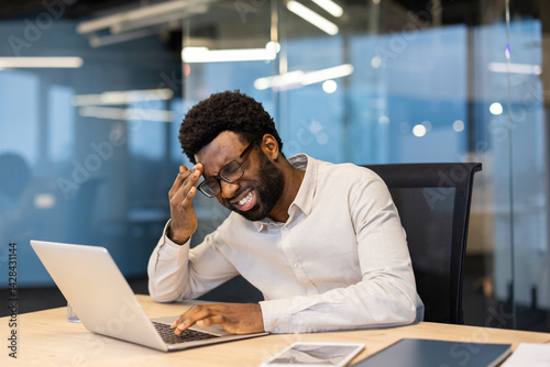 Obraz na płótnie An African-American man working at his desk is shown with a stressed expression. He is experiencing a headache and appears overwhelmed while using his laptop. w salonie